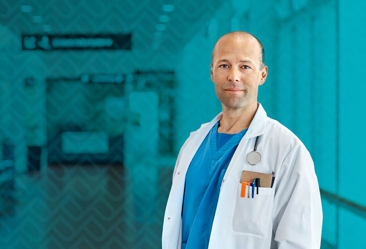 A male doctor looks toward the camera while standing in a hospital hallway.