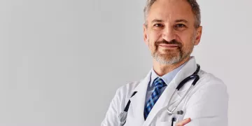 A physician stands with his arms crossed in front of a blank background.