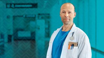 A doctor in blue scrubs and a white coat looks toward the camera while standing in the hallway of a hospital.
